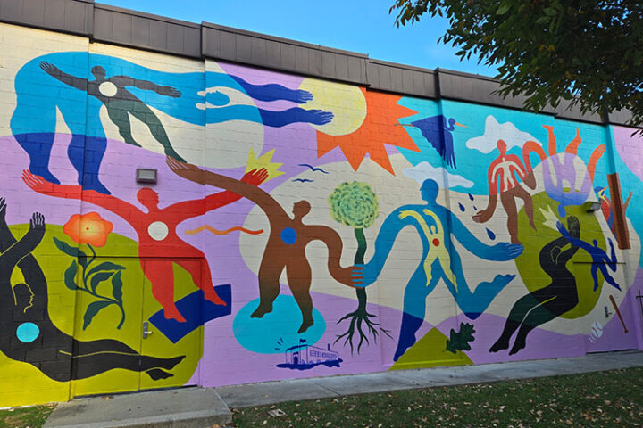 Colorful mural depicting abstract human figures, plants, and a sun on an exterior wall, with a tree partly visible in the foreground.