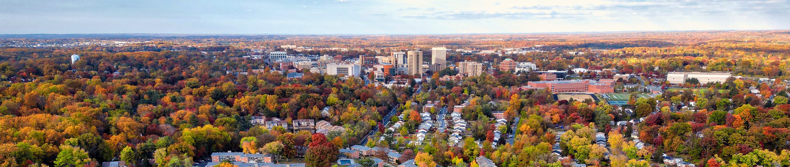 Aerial view of a city surrounded by autumn trees, with clusters of buildings and residential houses visible under a partly cloudy sky.