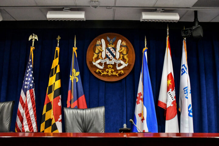 A row of empty chairs sits in front of six flags and a large official emblem mounted on a blue curtain backdrop in a formal meeting room.